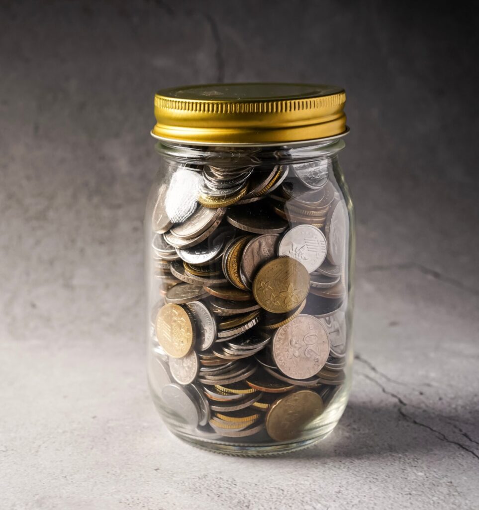 A jar of coins with the lid screwed shut, against a grey background. This represents someone's savings pot. Image by Adobe Stock.