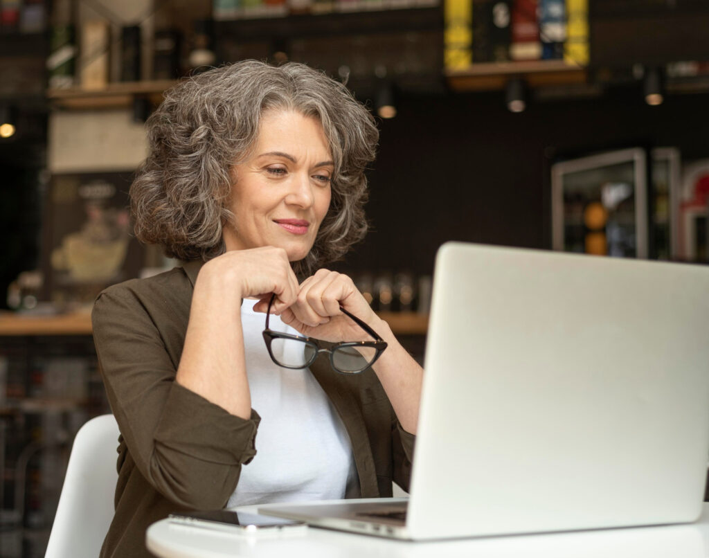 A middle-aged lady in a white t-shirt and brown cardigan looks at her laptop. Image by Freepik.
