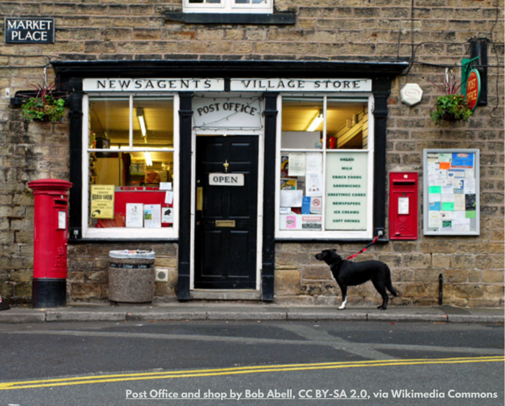 A photograph of a traditional village post office and shop. There are two red post boxes, one free-standing, one in the wall. A black and white dog is waiting for their person to come out of the post office.