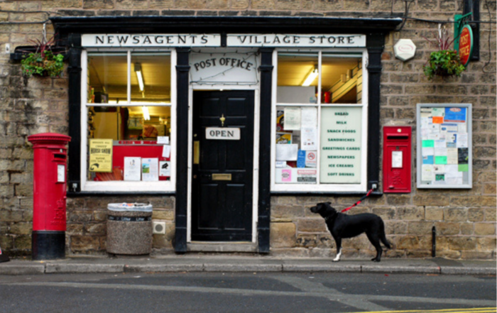 A photograph of a traditional village post office and shop. There are two red post boxes, one free-standing, one in the wall. A black and white dog is waiting for their person to come out of the post office.