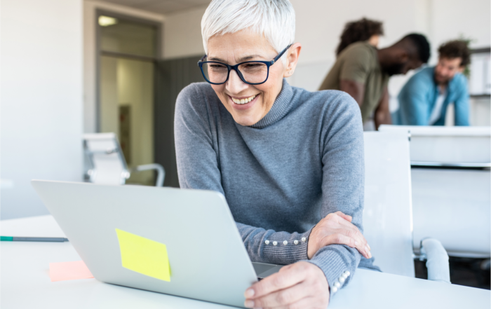 A smart grey haired, middle-aged woman smiles as she looks at her laptop. Image is © Adobe Stock.