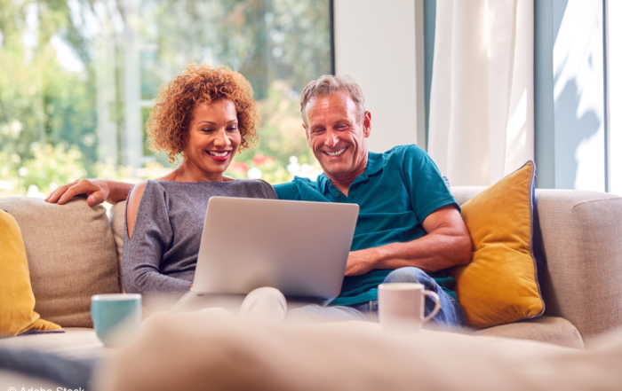 A middle-age couple are sitting on a couch with mugs of coffee smiling at their open laptop