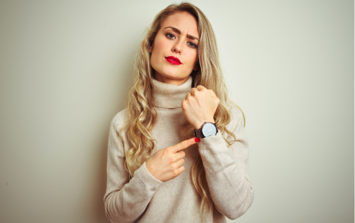 A woman in her 30s with long blonde hair and wearing a beige rollneck jumper. She is pointing at the watch on her left wrist, and looking impatient as though she has been waiting for too long. © Adobe Stock