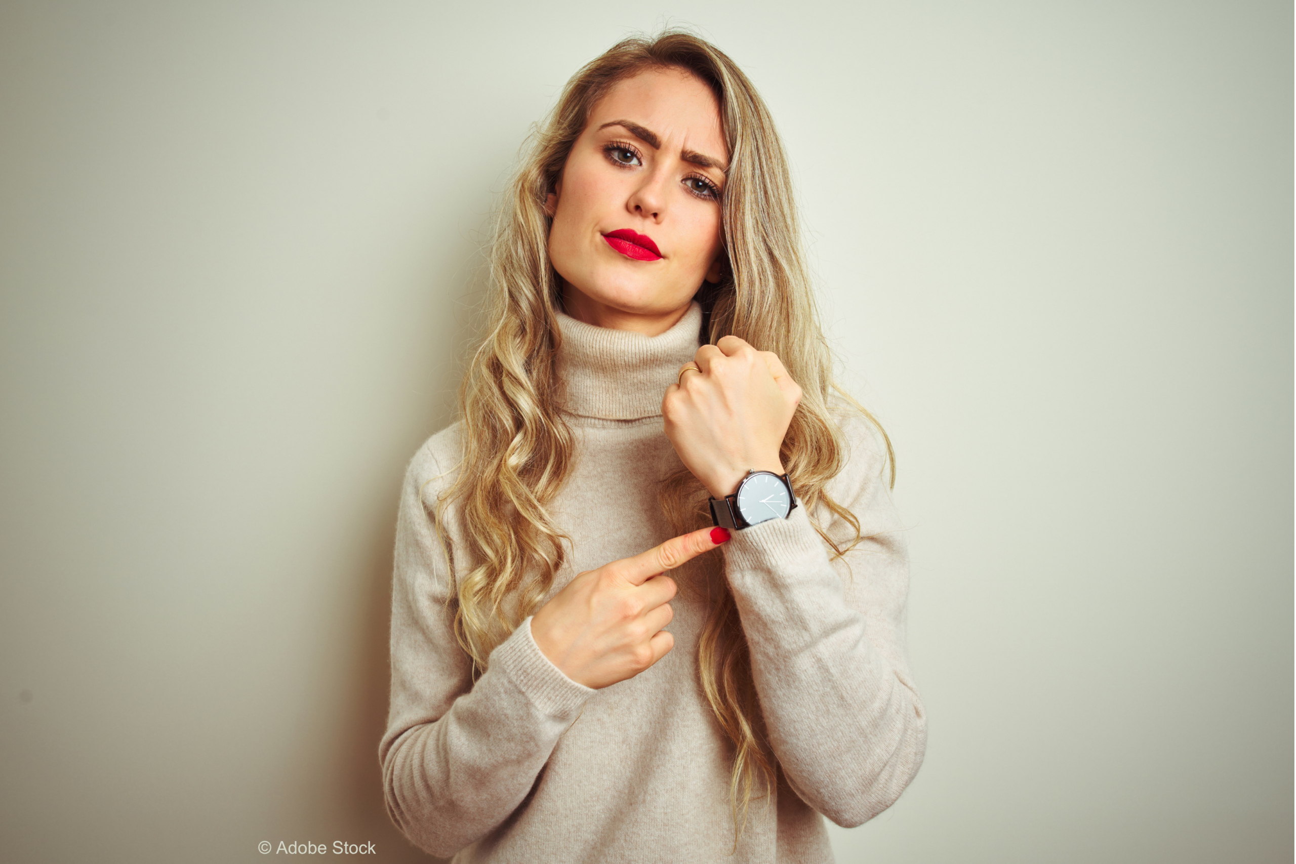 A woman in her 30s with long blonde hair and wearing a beige rollneck jumper. She is pointing at the watch on her left wrist, and looking impatient as though she has been waiting for too long. © Adobe Stock
