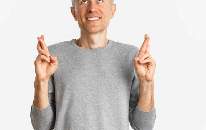 A man in his forties wearing a grey long-sleeved shirt has his fingers crossed. He is looking upwards, as if hopeful that good news is on the way.